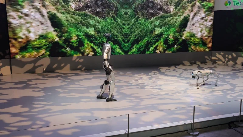 A silver and black Unitree G1 humanoid in side profile, with a Go2 quadruped to its right, on a lit exhibition stage in Tokyo.